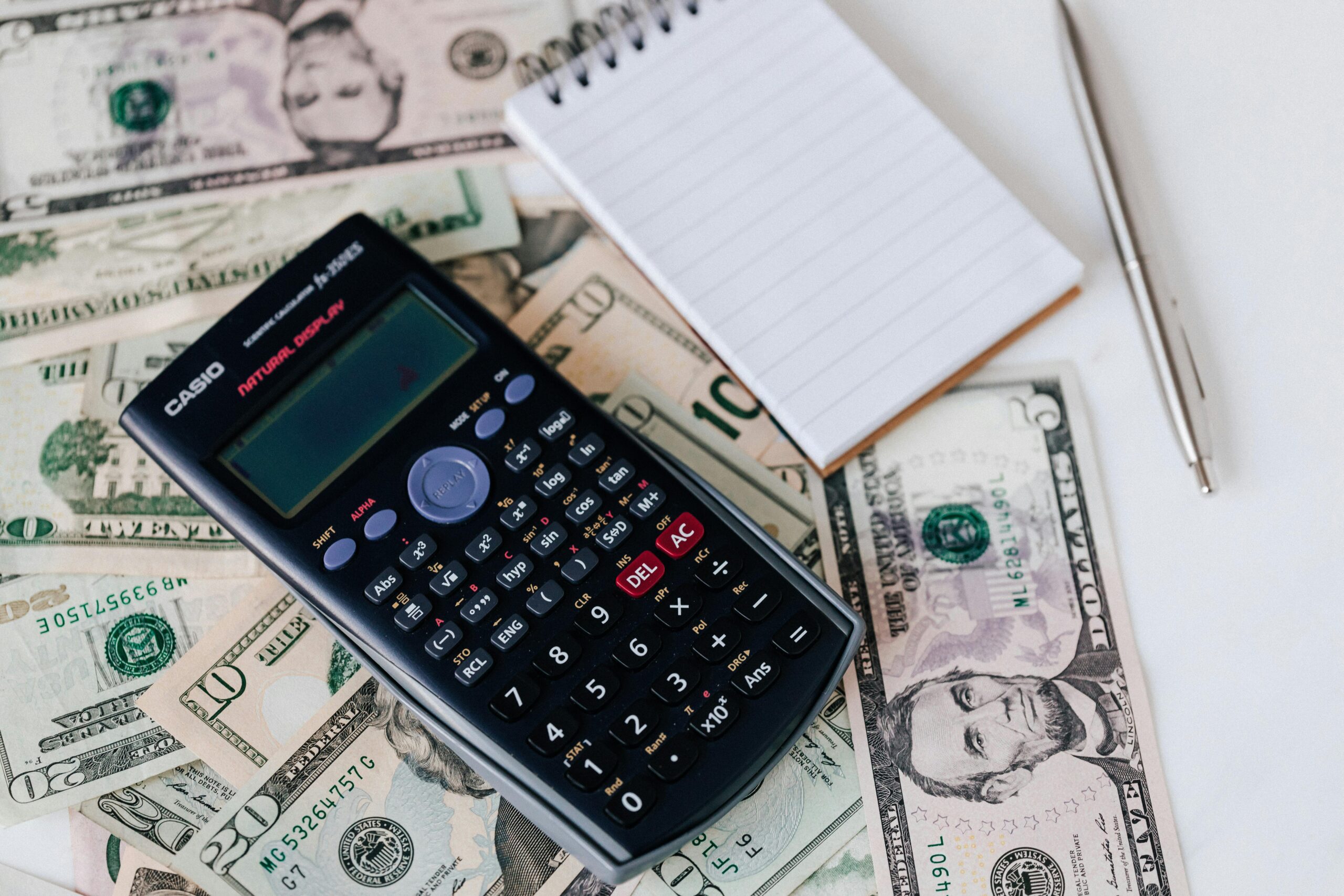 From above electronic calculator and notepad placed over United States dollar bills together with metallic pen for budget planning and calculation representing bookkeeping salisbury nc